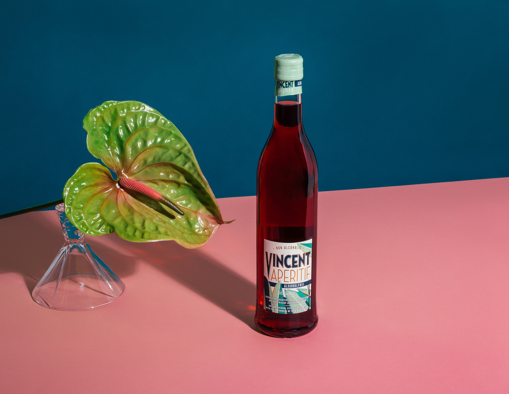 Eine Flasche Vincent Aperitif Alkoholfrei, in Szene gesetzt mit blau- und rosafarbenen Elementen